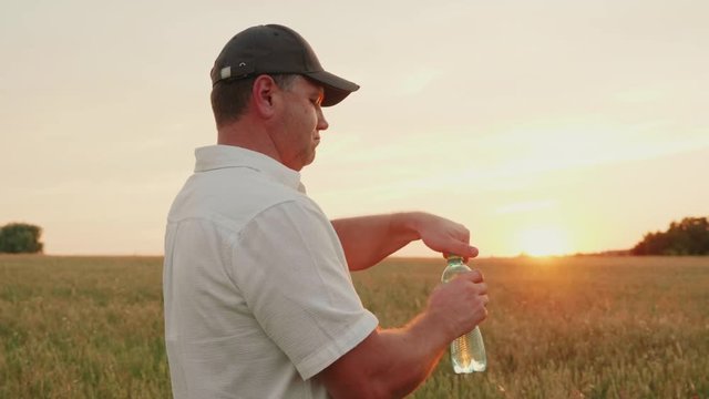 A middle-aged farmer man opens a bottle of water and drinks. Standing in the wheat field at sunset