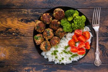 Fried meatballs with boiled white rice and broccoli on a black plate on the wooden rustic table, top view.