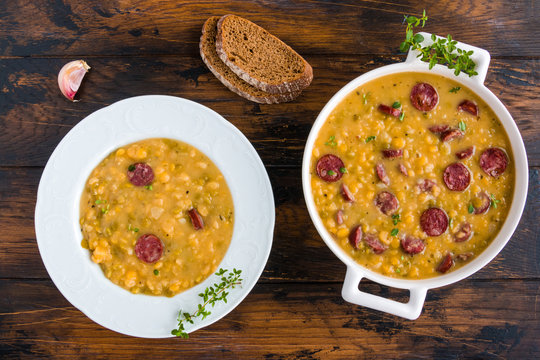 Hearty and healthy split pea and smoked sausage soup with green thyme in a white plate and casserole on the wooden table, top view.