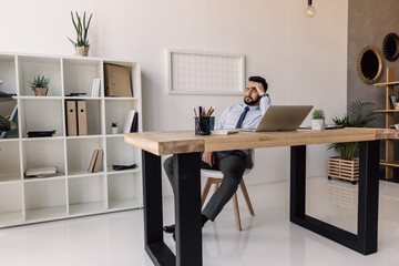 pensive bearded businessman sitting at table with laptop in modern office