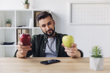 Smiling bearded businessman choosing between green and red apples while sitting at office table