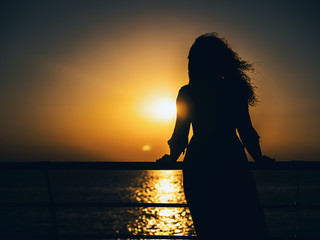 Silhouette of the happy woman in summer dress standing at the beach during sunset. Natural light and colors