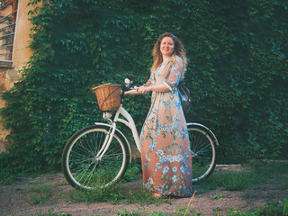 Beautiful young attractive woman standing near white retro bicycle on green wall with leaves background.