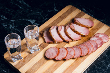 Shot glasses with vodka, slices of smoked meat and smoked sausage on wooden cutting board.