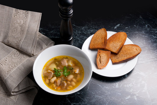 Bean soup in white plate, several toast on white plate on a black stone background.