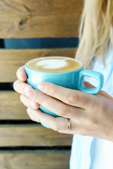 Closeup view on blue cup of cappuccino holding by young girl in jeans wear. Wooden background