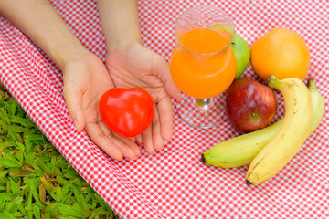 Close up of a young woman, holding a red heart, on the picnic mat with fruits. Healthy concept.