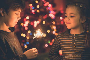 Young siblings, boy and girl looking at burning sparklers in the christmas eve evening with blurred christmas tree in background