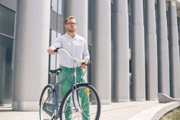 Cool young bearded hipster with bicycle on the city street. Smiling casual businessman having a break. He is standing in front of the building next to bike and holding laptop.