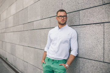 Cheerful young handsome man in sunglasses keeping hands in pockets and looking away with smile while standing against grey background. Stylish concentrated man.