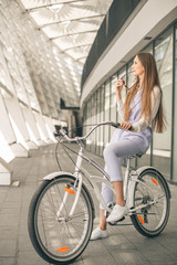 Fototapeta premium Woman with vintage bike. Side view of cheerful young woman in sunglasses and looking away while standing on her bicycle against the business center.