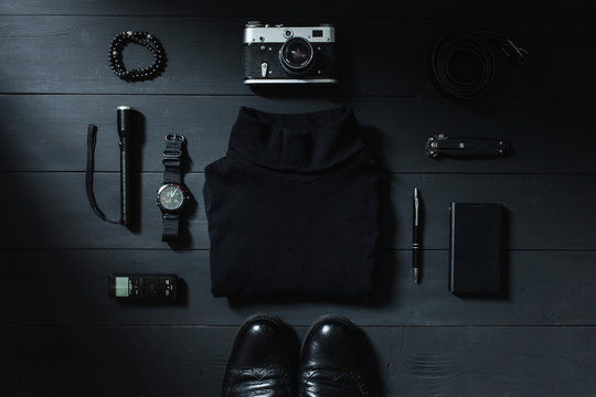 .On a dark table black things: a camera, a voice recorder, a knife, bracelet, turtleneck, belt, stick, flashlight, notebook, shoes, smart phone. Photographed from above. Male set..