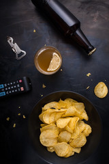 Plate of chips, remote control TV, beer, bottle opener top view of a dark metal table.