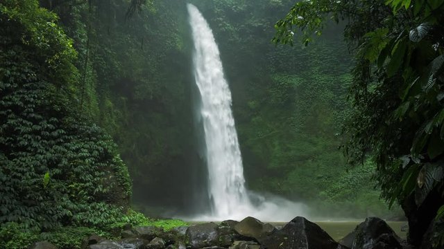 Amazing Nungnung waterfall, slow motioned falling water hitting water surface, some huge rocks seeable in front of frame. Lush green leafes is moving from the wind, Bali, Indonesia