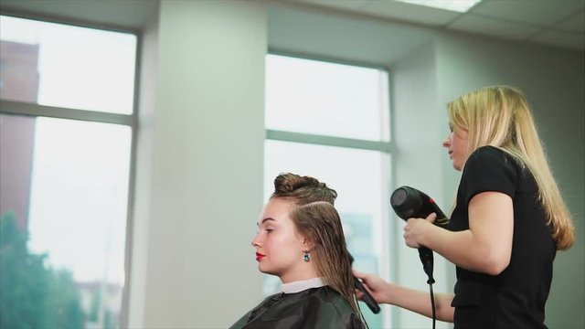 The blonde hairdresser dries long hair with the help of a warm air dryer and comb, a woman who came to the beauty salon expects a change of image and good service