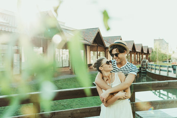 happy casual couple hugging while standing at bridge near cottage buildings, tree leaves on foreground