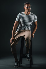 Handsome stylish man in sunglasses sitting on stool and looking away in studio