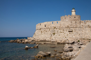 Fototapeta na wymiar Santa Claus Fortress on the pier in Mandraki Harbor. Bastion of defense on the quay of Rhodes. A defensive building in a harbor in the town of Rhodes with a lighthouse.