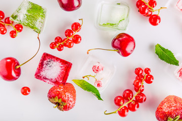Top view ice cubes with fresh berries among not frozen cherry, strawberry and mint leafs on the white background. Selective focus