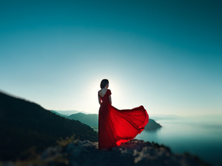 Young beautiful woman in red dress looking to mountains sea - obrazy, fototapety, plakaty