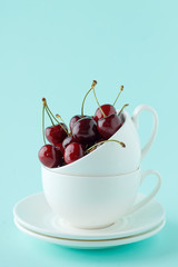 Stack of two white teacup with sweet ripe cherry on pastel cyan background.