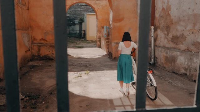 Young attractive woman wearing long skirt walking near vintage white bike in old european courtyard. Girl returns home. Slow motion.