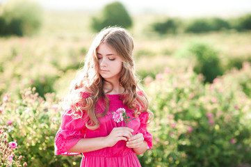 Fototapeta premium Beautiful blonde teen girl 14-16 year old wearing pink dress holding rose flower outdoors. Summer portrait.