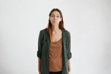 Adorable young female with appealing appearance puffing out her cheeks while standing over white...