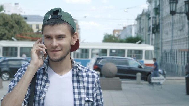 Young handsome man in cap walking in the street, talking on the phone. Steadycam shot. People stock footage shot at summer season time. 