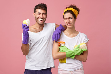 Smiling man wearing protective gloves and white T-shirt holding sponge in hand leaning at his pretty wife who is dissatisfied not wanting to do cleaning standing crossed hands having frowning face