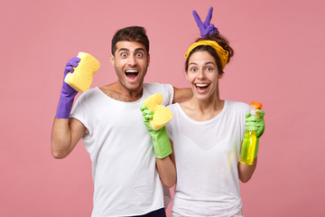 Young male and female playing fools while standing against white pink wall resting after doing cleaning in their appartment holding sponges and spray laughing and looking with excited faces in camera