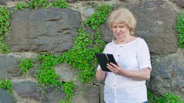 Elderly woman using tablet computer with earphones. Vintage wall of wild stone in the background.