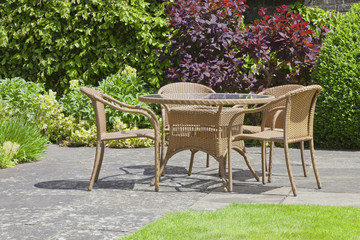 Rattan dining chairs and table in a garden patio surrounded by plants and flowers, on a sunny summer day .