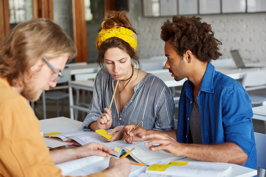 Portrait Of College Students From Different Countries Meeting Together Sharing Their Experience Helping Each Other With Studying Having Serious And Concentrated Looks In Books Sitting At Table Indoors