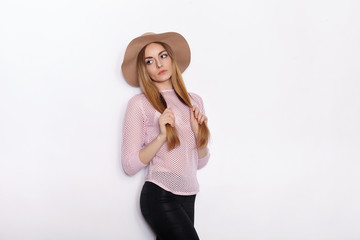Cute and stylish beautiful young blonde woman in trendy clothing practicing model poses and looking away with smile while standing against white wall background