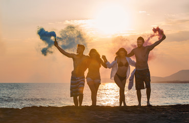 Group of happy people walkin on beautiful beach in summer sunset in their hands is smoke bomb