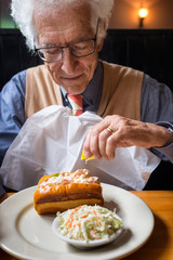 Happy senior man eating at a restaurant squeezes lemon onto his seafood sandwich. Lobster rolls are a regional summer specialty in Maine, USA. - 162772525