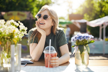 Portrait of young adorable woman drinking lemonade in park surronded by flowes, wearing glasses dreaming of her future career as a beaty blogger.