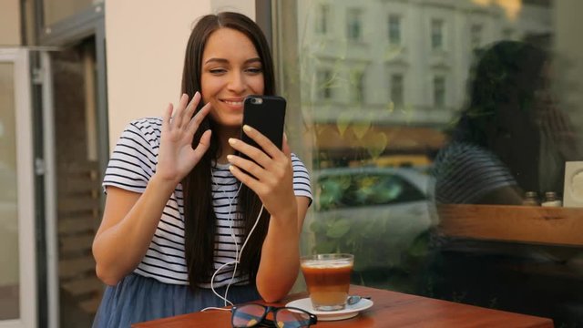 Attractive young woman video cahtting, skyping with her smart phone. Woman waving at the camera while sitting outdoors inh the cafe
