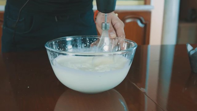 Backer unrecognisable man mixing milk in glass bowl using whisk on kitchen table, close up dolly shot