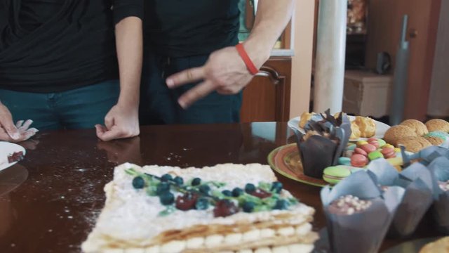 Pastry cooks caucasian couple posing, smiling and makes hand signs over table with desserts cakes, muffins, macarons at kitchen