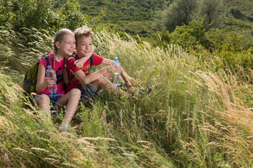 Boy and girl are sitting in high grass and holding water bottles