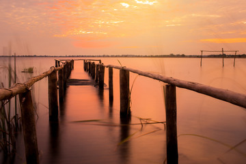 Fototapeta na wymiar long exposure of wooden old bridge into the lake on the background of worm sky sunset.