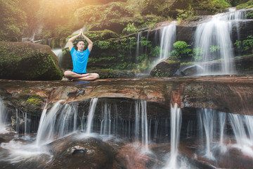 Young man meditating in lotus position while doing yoga on the rock between waterfalls