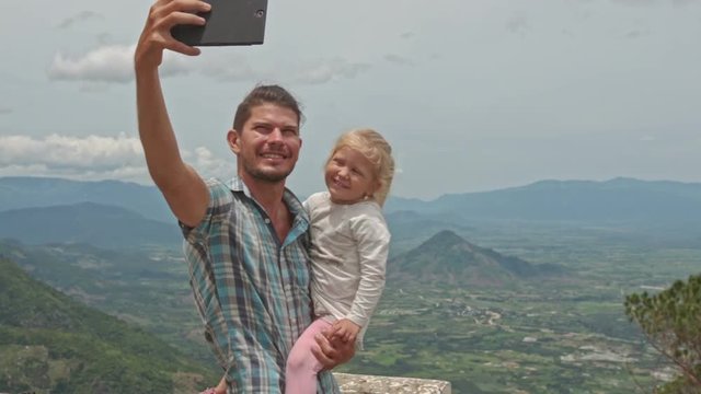 closeup caucasian father holds in arms little blond daughter makes selfie girl kisses to cheek against green valley