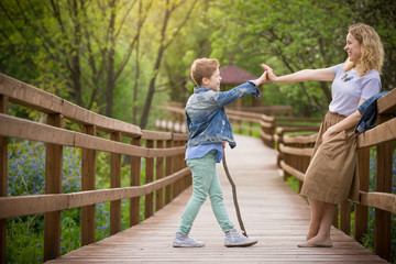 Obraz premium Portrait of young blond woman standing on a wooden bridge and giving five to her cute son in the park on a sunny summer day. Lifestyle. Outdoor.