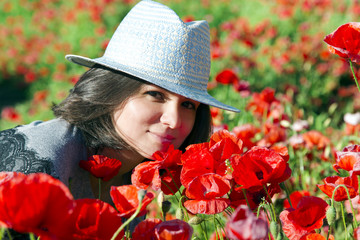 Portrait of beautiful brunette in a summer hat against a background of red poppies in the height of summer.Beautiful woman enjoying the bright red wild flowers, harmony concept.
