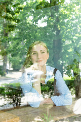 Behind the glass portrait of young and attractive blonde girl sitting in cafe with a cup of aroma cappuccino