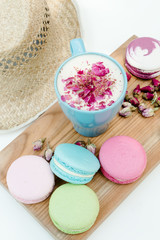 Obraz na płótnie Canvas Tasty macaron cookies with a blue cup of cappuccino with roses petals and straw hat on white table.