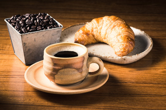 espresso cup on wooden table with butter croissant in dish and coffee bean bucket at back